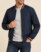 A man wearing a navy blue bomber jacket with red and white striped ribbed collar, cuffs, and hem, styled over a light gray t-shirt and dark blue jeans against a solid beige background.