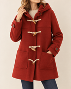 A woman wearing a red hooded duffle coat with cream rope toggle closures and large front patch pockets, paired with a cream ribbed sweater and blue jeans, against a plain light beige studio background.