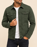 A man wearing an olive green button-up denim jacket over a white t-shirt and dark jeans, shown from the chin down against a solid beige background.