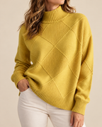 A woman wearing a mustard yellow ribbed knit mock neck sweater with a diamond argyle pattern on the front, paired with white pants, against a solid beige background.