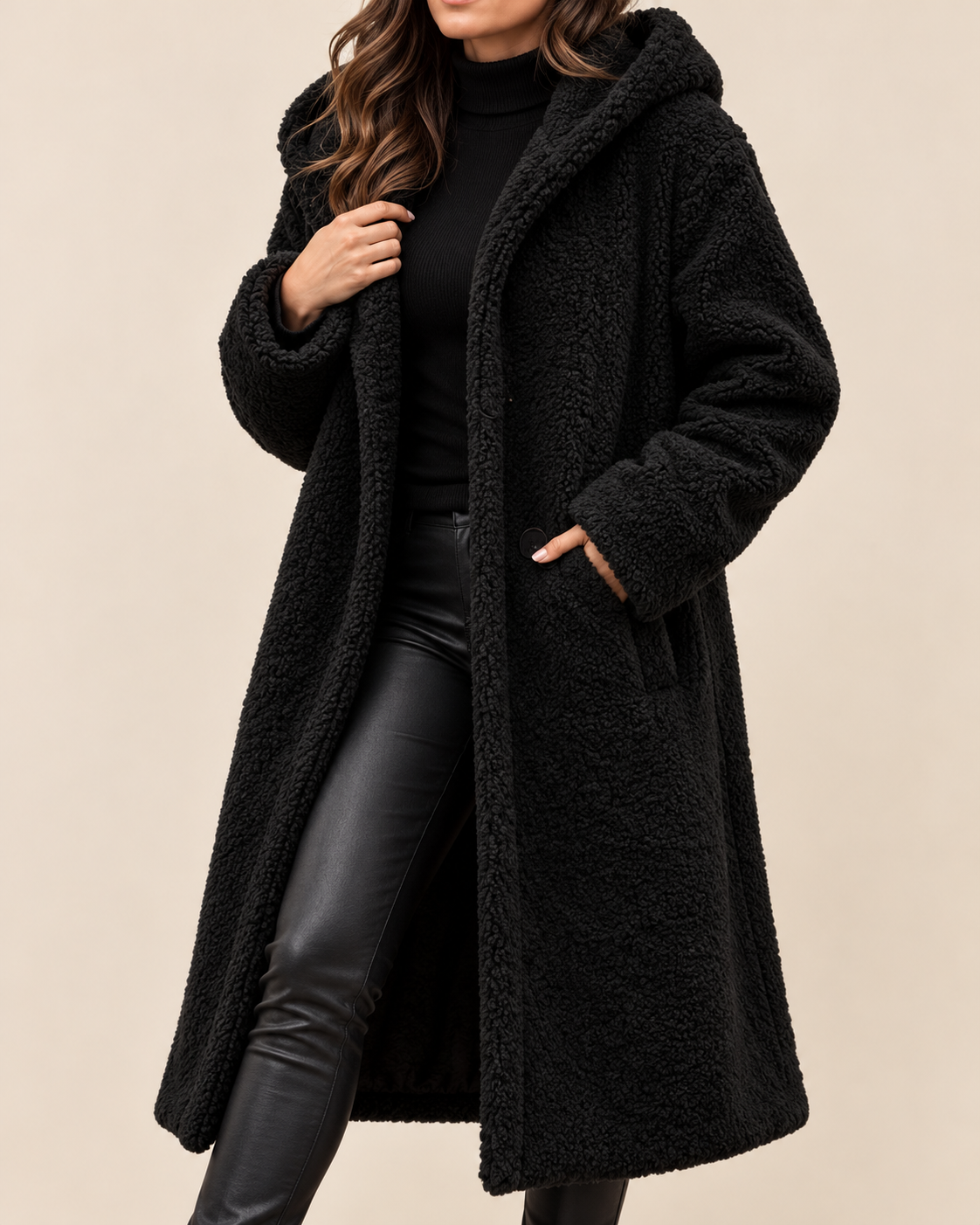 A woman wearing a long black teddy fleece hooded coat over a black turtleneck and black faux-leather pants, standing against a plain beige studio background.
