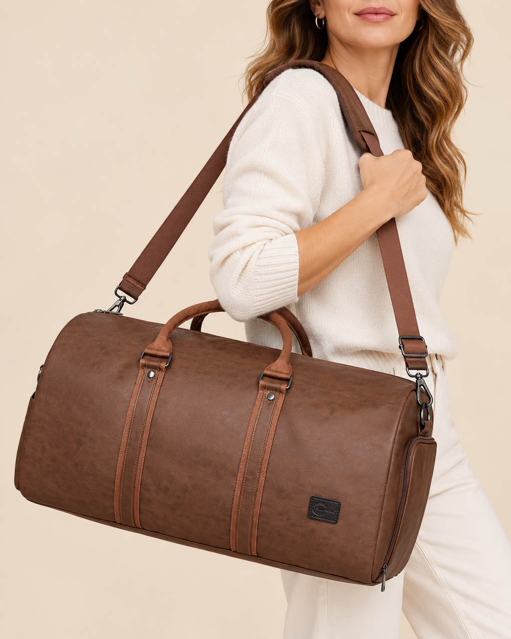 A woman wearing a cream knit sweater carries a brown faux-leather duffel bag with a matching shoulder strap against a solid beige background.