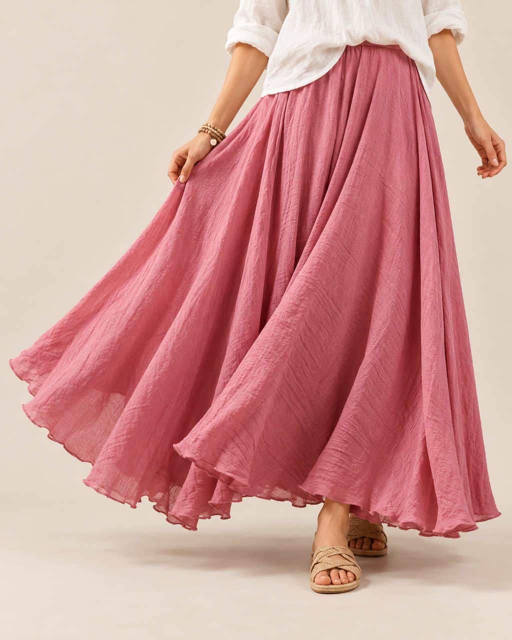 A woman wearing a flowing dusty pink maxi skirt with a white blouse and beige sandals, shown from the waist down against a plain beige studio background.