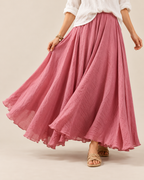 A woman wearing a flowing dusty pink maxi skirt with a white blouse and beige sandals, shown from the waist down against a plain beige studio background.