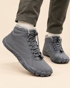 A pair of gray lace-up ankle boots with a fuzzy gray lining, shown on a model wearing dark olive pants and white socks against a light beige background.