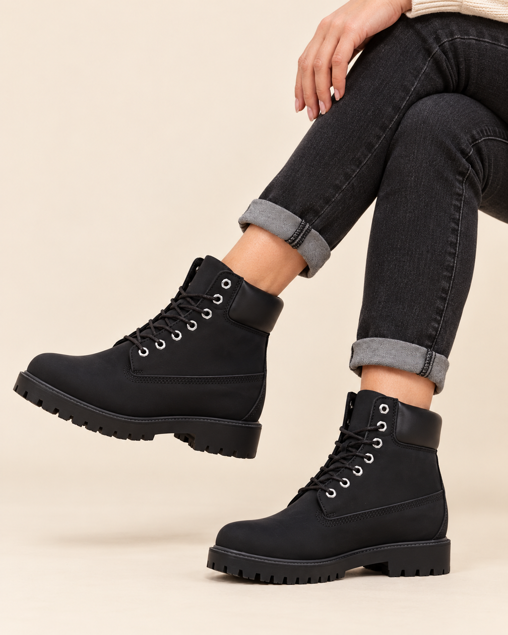 A person wearing black lace-up ankle boots with chunky lug soles and dark gray cuffed jeans, shown against a plain beige studio background.