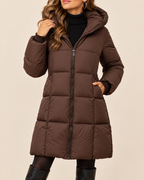 A woman wearing a brown hooded puffer coat over a black turtleneck sweater, paired with black tights and black boots, against a solid beige background.