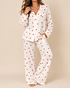 A woman modeling a two-piece white pajama set with a red heart pattern, consisting of a long-sleeve button-up top and matching wide-leg pants, set against a solid beige background.