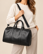 A woman wearing a cream knit sweater and light beige pants carries a black faux-leather duffel bag with top handles and a shoulder strap against a plain beige background.