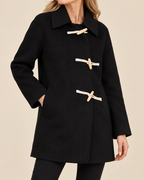 A woman wearing a black duffle coat with light beige toggle closures, shown from the neck down against a plain beige studio background.