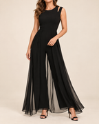 A woman modeling a sleeveless black jumpsuit with a high round neckline, fitted bodice, and wide flared legs with sheer overlay panels, shown against a plain beige studio background.