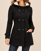 A woman wearing a black hooded duffle coat with a white sherpa-lined hood and dark toggle closures, paired with blue jeans, against a solid beige background.