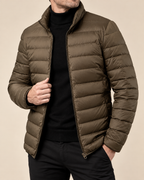 A man wearing a dark olive green puffer jacket over a black turtleneck sweater and black pants, shown from the chin down against a plain beige background.